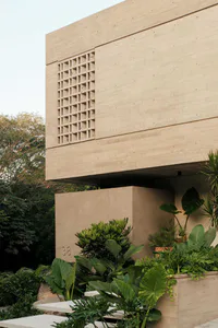Exterior view of a modern concrete house with a decorative breeze-block wall and abundant tropical plants in the foreground.