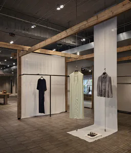 現代極簡精品店室內空間，粗獷原木天花板大樑、白色薄紗織物隔間，以及三件懸掛展示的設計師服裝：黑色洋裝、淡黃色無袖長袍與灰色中式立領外套。
