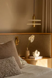 Close-up of a modern bedroom corner showing a wooden nightstand, beige bouclé pillows, a wall-mounted brass reading light, a hanging two-tier brass and alabaster pendant lamp, and decorative objects including a ceramic vase with a white anthurium flower.