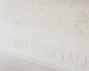 Close-up view of a white marble wall featuring raised, carved lettering that reads 'CAFFÈ SOCIALE' in an elegant serif font, with a speckled terrazzo floor visible below.