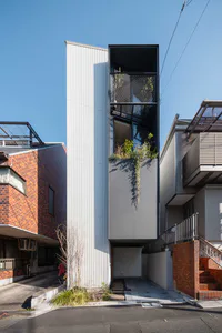 高聳狹窄的現代町屋，外牆為白色波紋金屬板，深色金屬網格陽台種滿植物，垂掛綠意對比晴朗藍天，兩側為鄰近的磚造與混凝土建築。
