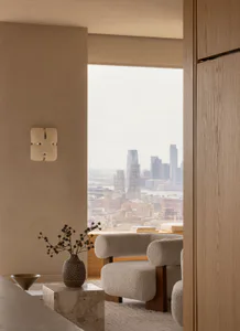 Modern minimalist living room interior with light oak wood paneling, textured boucle armchairs, travertine side table, dried floral arrangement, and a large window overlooking the New York City skyline across the Hudson River.