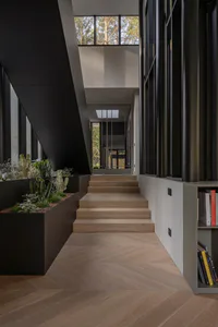 Modern minimalist interior entryway with light oak herringbone floor, wide oak staircase, large black concrete planter filled with cacti and succulents, dark metal and glass walls, and high clerestory windows overlooking pine trees.