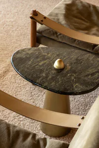 Close-up view of a round green marble side table with a brass knob on a beige carpet, positioned next to a modern armchair with tan leather straps and olive green upholstery.