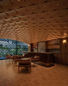 Elegant modern hotel bar interior with dark walnut bar counter, brown leather armchairs, round wooden tables, and a striking coffered wooden ceiling with recessed lighting, next to a large patterned window overlooking greenery.