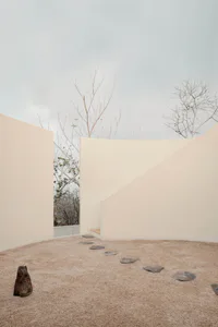 Minimalist outdoor courtyard with beige curved walls, a sandy ground, irregular dark stepping stones, a small wooden bench, and bare trees against an overcast sky.