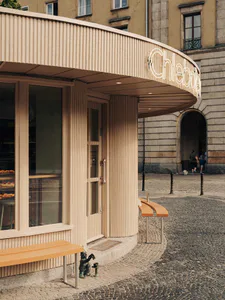 Curved beige bakery storefront with vertical fluted cladding, glass display windows showing pastries, wooden benches, and a small bronze statue on a cobblestone square in Warsaw.