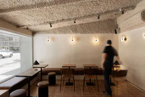 Interior view of a modern minimalist cafe with a heavily textured plaster ceiling, terracotta floor tiles, wooden tables, built-in banquette seating, and a person walking through the space.