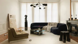 Modern Parisian living room featuring a large curved navy blue velvet sofa with a beige fur throw, a sculptural black chandelier, minimalist metallic coffee table, olive-green armchair, abstract painting, and dark wood fireplace mantel.