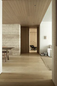 Modern minimalist interior hallway with a large textured travertine stone wall on the left, light oak wood plank ceiling and flooring, a wooden dining table, and a view into a living area with a black leather Eames lounge chair and white sofa.