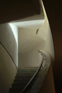 View looking down a curved concrete spiral staircase with a smooth handrail and dramatic natural light creating strong contrasts on the walls.