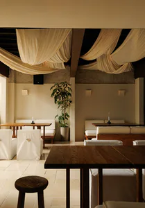 Modern minimalist restaurant interior featuring wooden tables, white upholstered seating, a tall potted plant, and dramatic draped cream fabric across the ceiling.