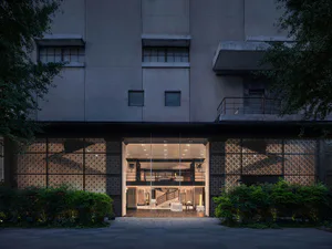 Nighttime exterior view of a modern concrete building with large glass entrance, intricate patterned lattice screens illuminated from within, and lush greenery on both sides.