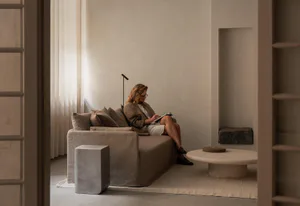A woman with blonde hair and glasses sits on a beige sofa writing in a notebook in a minimalist living room featuring a round travertine coffee table, concrete side table, and neutral-toned walls and curtains.
