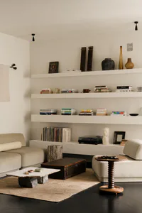 Modern minimalist living room with white walls, built-in floating shelves displaying books and decor, a travertine coffee table on a jute rug, a vintage trunk, beige sectional sofa, and a wooden side table.