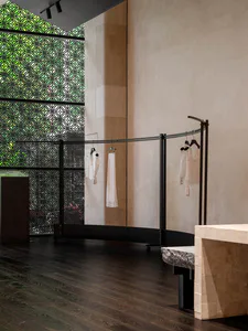 Interior view of a minimalist boutique featuring a curved black metal clothing rack with white lace and embroidered garments, a large travertine display block in the foreground, dark wood flooring, and a decorative green patterned window screen in the background.