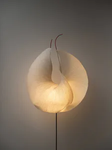 A modern sculptural floor lamp featuring a large, glowing, pleated white paper shade shaped like a twisted torus, mounted on a thin black metal pole against a neutral gray background. Two thin red wires emerge from the top of the shade.