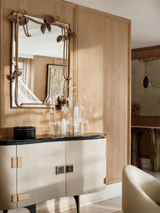 Elegant entryway featuring a curved beige console table with black marble top, paired with an ornate brass mirror decorated with botanical motifs, set against light oak wood paneling.