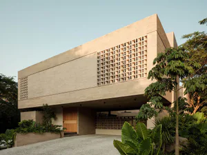 Exterior view of a brutalist-style concrete house with geometric lattice screens, wooden entrance door, and lush tropical landscaping under a clear sky.
