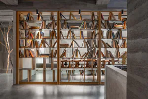 Modern concrete interior featuring a large double-sided oak bookshelf filled with books, wooden reading tables and stools visible through the shelves, with concrete walls and polished floor.