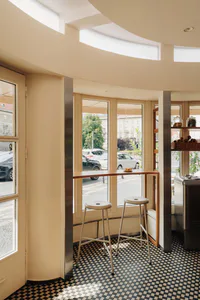 Bright cafe interior featuring a curved wooden breakfast bar, two minimalist stools, black and white checkerboard floor tiles, and large arched windows overlooking a European street.