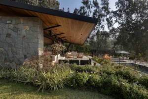 Modern outdoor patio featuring a large stone wall, extensive wood-paneled ceiling overhang, contemporary outdoor furniture, and a lush garden with swimming pool in the background.