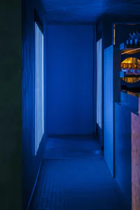 Narrow concrete corridor illuminated by intense blue vertical LED light strips on both walls, leading to a modern bar area with warm backlighting and glassware display.