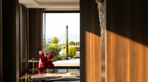 Modern luxury living room interior featuring a vibrant red flower-shaped armchair, dark walnut wood paneling, glossy reflective floor, and large glass doors opening to a landscaped terrace with mountain views.