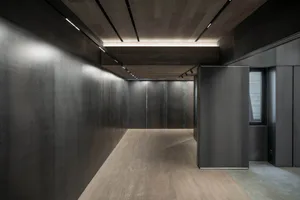 Modern minimalist corridor with dark metal or concrete panel walls, light wood flooring, and recessed linear ceiling lights creating a dramatic atmosphere.