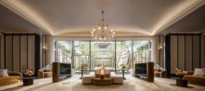 Elegant luxury hotel living room with a large crystal chandelier, curved sofas in warm tones, two wall-mounted TVs, and a view of a traditional Chinese garden through floor-to-ceiling windows.