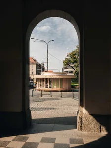 View through a massive stone archway looking out onto a sunny European square with a light-colored circular wooden kiosk and historic buildings in the background.