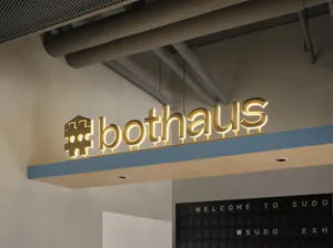 Illuminated golden brass 3D sign spelling 'bothaus' with a stylized hashtag logo mounted on a blue and wood architectural canopy in a contemporary office lobby, with a black letter board below reading 'WELCOME TO SUDO #SUDO EXH'.