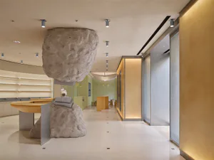 現代零售空間內部，一塊巨大的天然清水模巨石懸浮在弧形淺木色櫃檯上方，下方有較小巨石基座，背景為淡綠色牆面與溫暖環境照明。