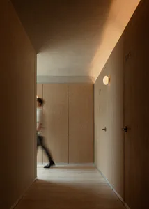 A long, minimalist hotel hallway with light oak wood paneling on walls and doors, round wall sconces emitting warm light, and a person captured in motion blur walking from a connecting corridor.