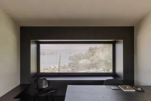 Minimalist home office interior featuring a large horizontal window with blinds, a built-in dark wood window seat, a black molded chair, and a concrete-like desk with minimal accessories.