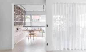 View into a bright minimalist home office featuring light wood built-in cabinetry, a simple wooden desk and chair, large windows with roller blinds, and white sheer curtains on the right.