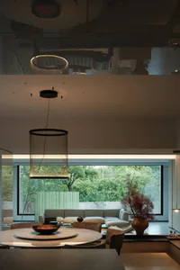 Modern open-plan dining and living room featuring a large round travertine table, woven string pendant light, beige sectional sofa, and floor-to-ceiling windows overlooking a lush green garden.
