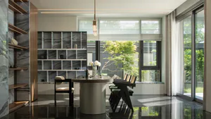 Modern dining room featuring a cylindrical marble pedestal round table, dark wood dining chairs with green upholstery, built-in marble shelving unit, and large windows overlooking a lush garden.