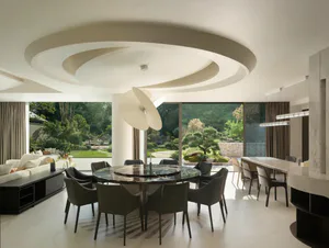 Modern luxury dining room with a large round dark marble table surrounded by eight dark green upholstered chairs, a sculptural white pendant light hanging from a circular coffered ceiling, and large glass doors opening to a lush Japanese-inspired garden.