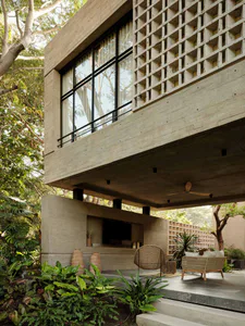 Modern tropical house exterior featuring exposed concrete structure, large black-framed windows, built-in entertainment niche, and an open-air patio furnished with natural wicker lounge chairs and sofas surrounded by abundant tropical plants.