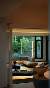 Modern minimalist living room interior featuring a beige linen sectional sofa, low black coffee tables, a woven daybed, and tall floor lamps, with large windows overlooking a dense green forest.
