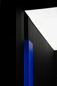 Low-angle view of a dark wooden architectural wall with a glowing bright blue vertical resin or acrylic column against an overcast sky.