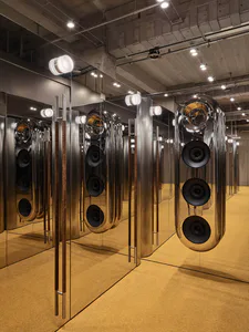 Large polished stainless steel cylindrical speakers with exposed drivers, suspended in a mirrored room with yellow carpet and industrial ceiling.