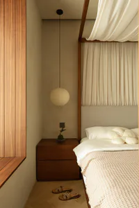 A minimalist bedroom corner with a walnut wood nightstand, Akari paper lantern pendant light, built-in wooden window seat, beige upholstered bed with canopy, and a small bonsai plant.