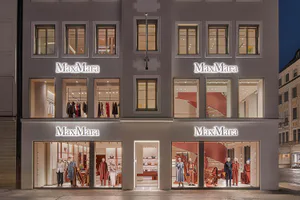 Night view of a MaxMara luxury fashion boutique with four illuminated ground-floor display windows featuring mannequins in autumn-winter collections, red spiral staircase visible inside, and glowing white MaxMara signage on a classical stone building facade.