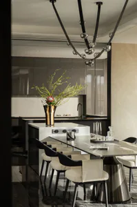 Modern luxury dining room with a long marble dining table, cream and black dining chairs, a white marble kitchen island with a large floral arrangement in a gold vase, and a striking modern black chandelier with multiple glass globes hanging above.