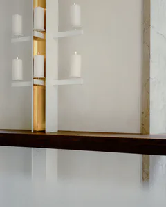 Modern minimalist wall installation with white pillar candles on white floating shelves, set against a recessed brass panel with warm backlighting, above a dark wood mantel and next to a white marble wall.