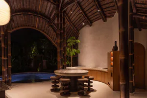 Night view of a luxurious outdoor kitchen featuring a large round marble dining island with wooden stools, set under an intricate vaulted bamboo ceiling with a glowing swimming pool visible in the background.