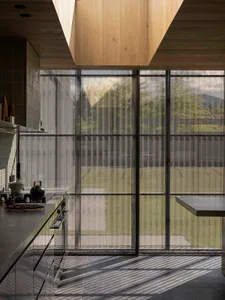 Modern kitchen interior featuring dark concrete countertops, stainless steel appliances, a black kettle on the stove, and large grid windows with sheer curtains overlooking a green landscape and mountains.