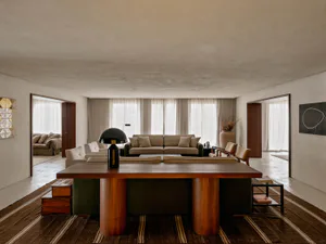 Spacious minimalist living room featuring a large round walnut dining table on a striped rug, neutral sofas, and large windows with sheer curtains.